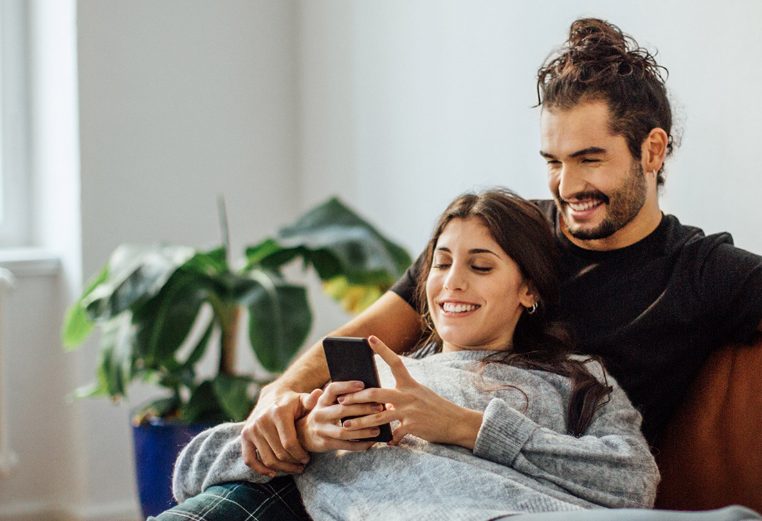 A man and woman sitting on a sofa together, smiling while looking at a cell phone.