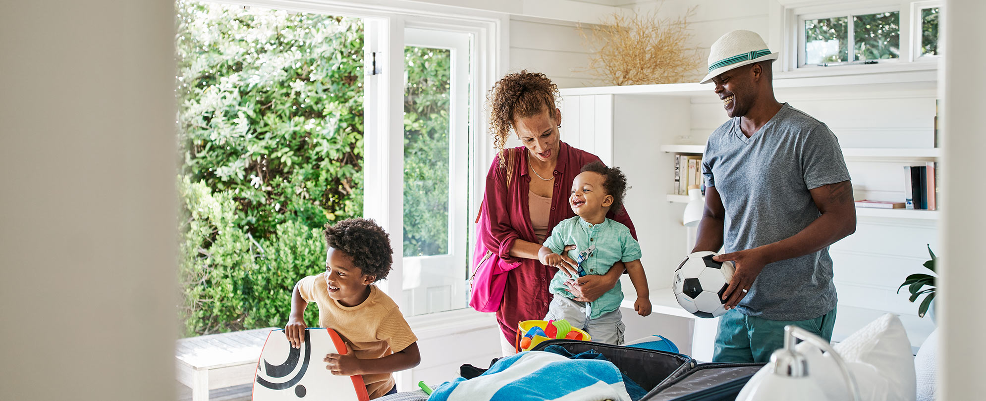 A smiling, family of four standing around a table, near an open window with greenery on the other side. 