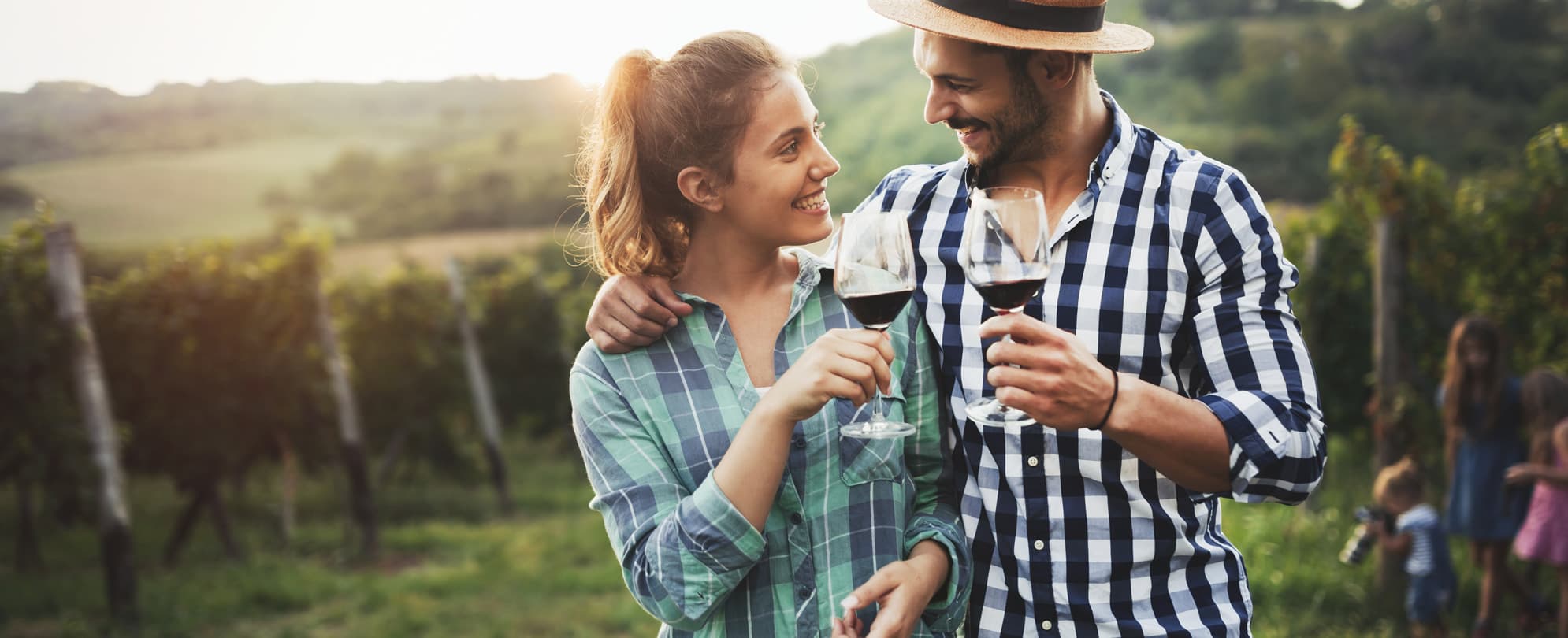 A couple in their 30s clink wine glasses full of red wine in a sunny vineyard