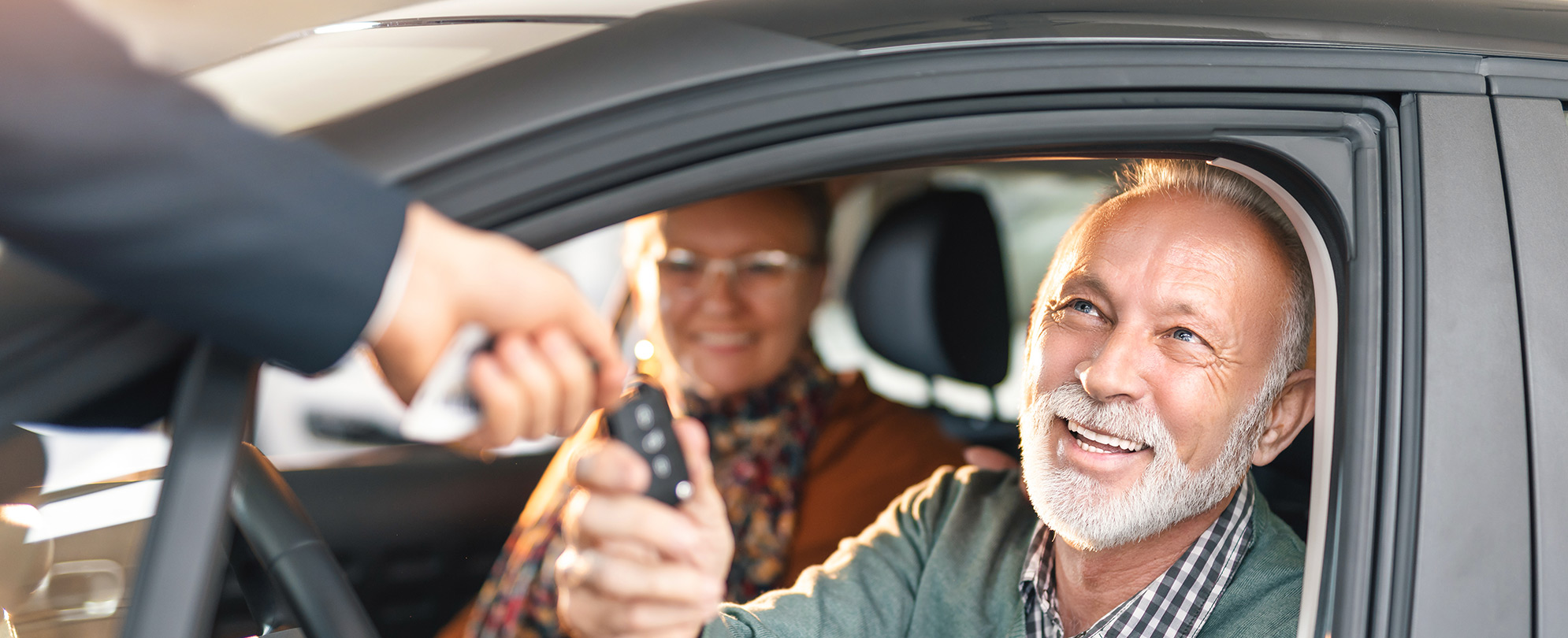 A man handing over car keys to a couple sitting in a car.