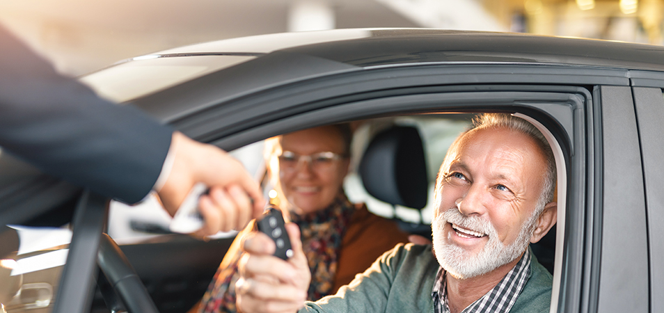 A man handing over car keys to a couple sitting in a car.