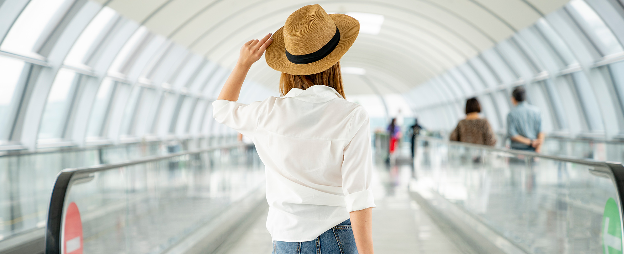 Woman holding her hat on her head and facing forward on a moving walkway at an airport.