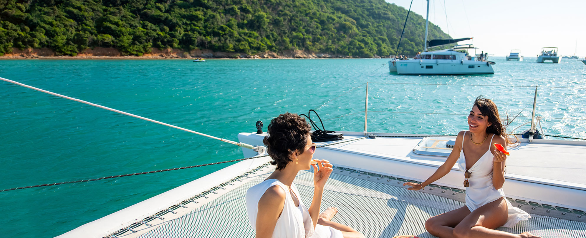 Two women sunbathing and eating watermelon on a catamaran boat.