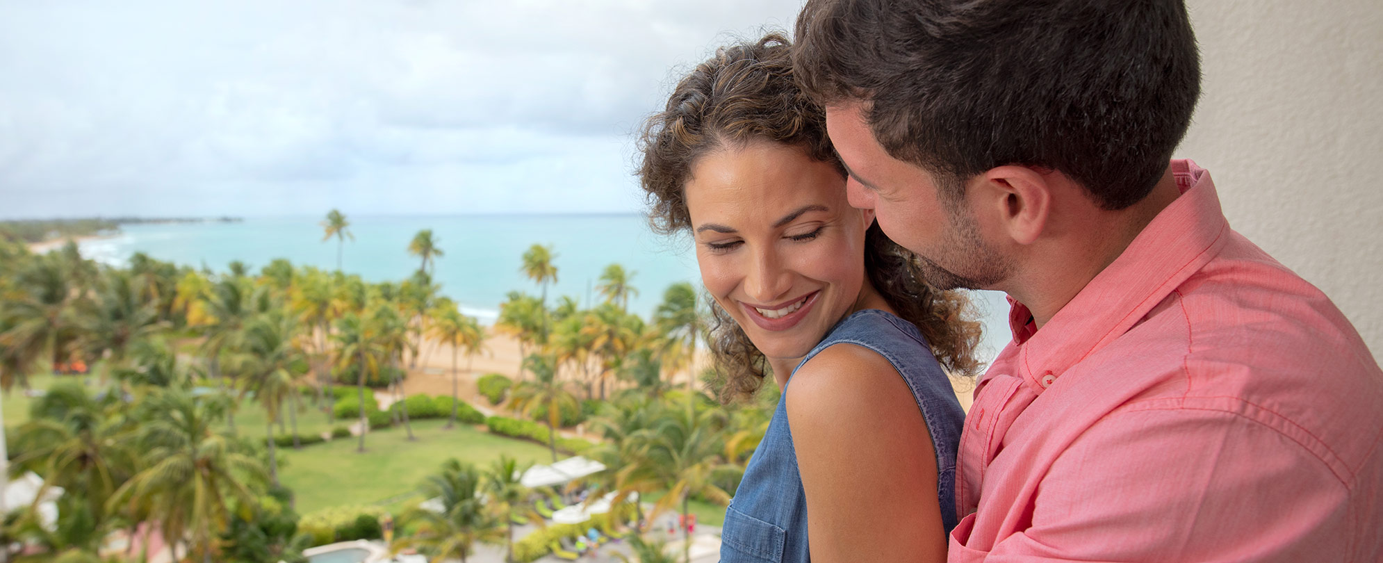 Man and woman smile on the balcony of their Margaritaville Vacation Club suite, with palm trees and ocean in the background.