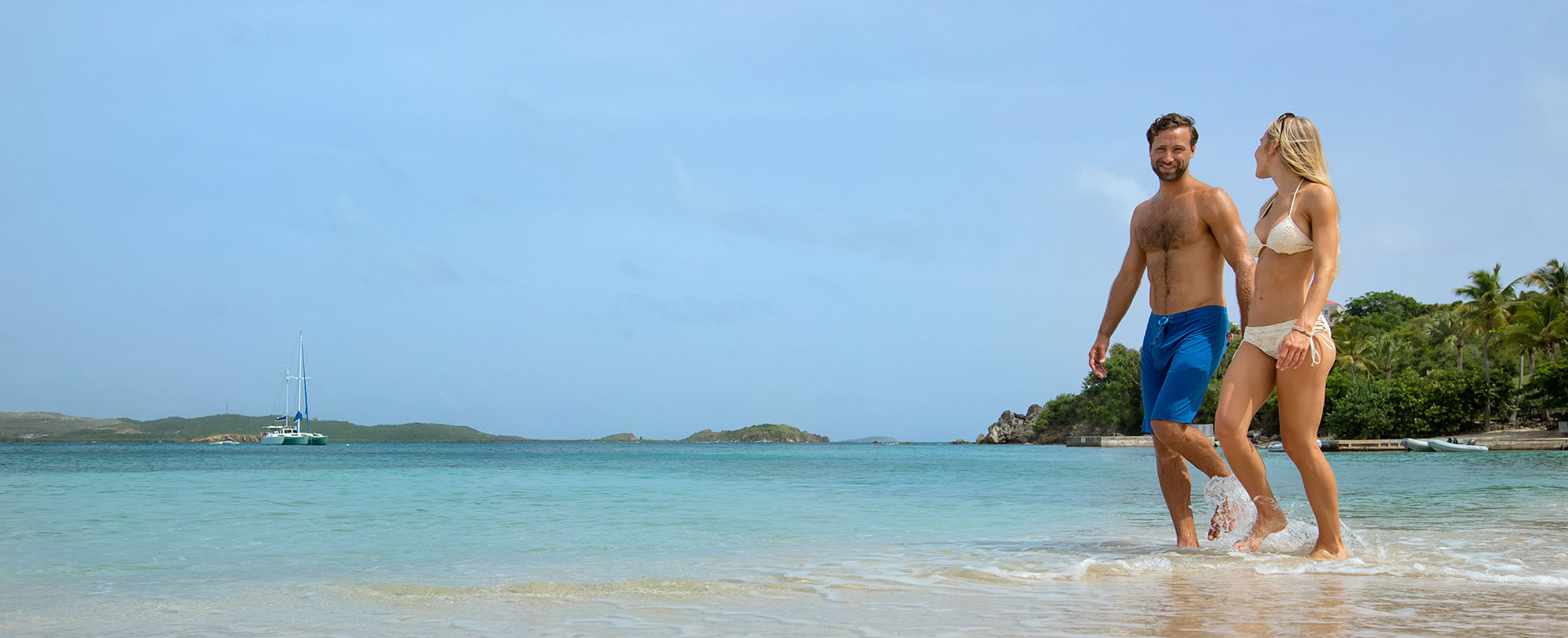 Man and woman wearing swimsuits walk through clear ocean water outside of a Margaritaville Vacation Club resort.