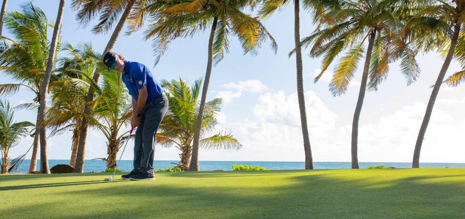 Man in a blue polo putting a golf ball at an oceanfront golf course near a Margaritaville Vacation Club resort.