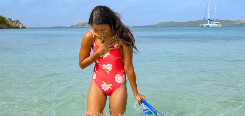 Young woman wearing a red, one-piece swimsuit, walks in the ocean holding mask and snorkel on a Margaritaville vacation.