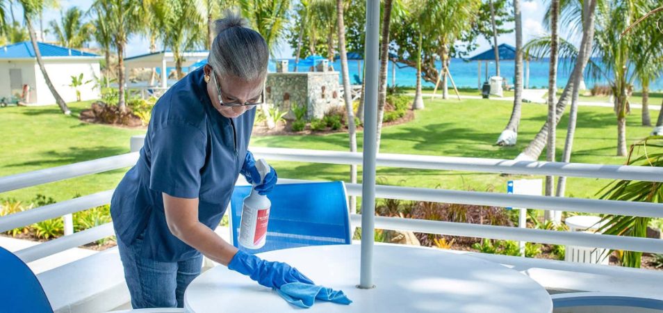 Female employee wearing a blue medical mask and gloves wipes an outdoor table at a Margaritaville Vacation Club resort. 