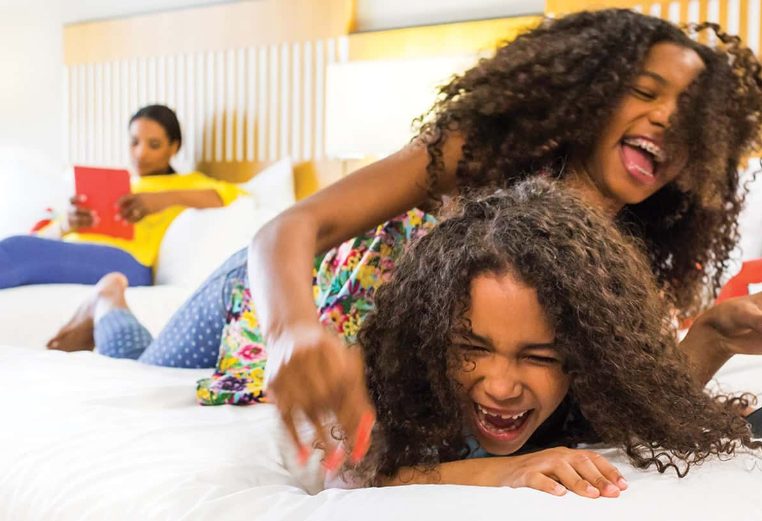 Two smiling young girls with dark, curly hair wrestle for remote on the bed of a Margaritaville Vacation Club Resort suite.