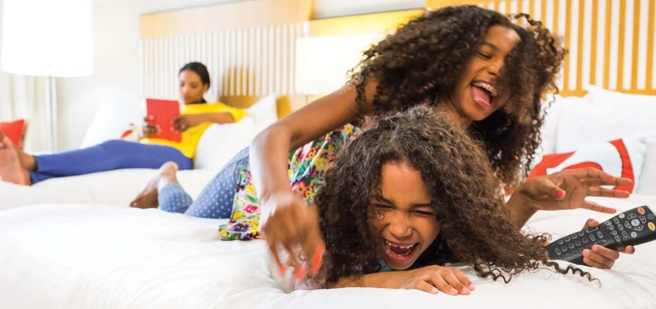 Two smiling young girls with dark, curly hair wrestle for remote on the bed of a Margaritaville Vacation Club Resort suite.