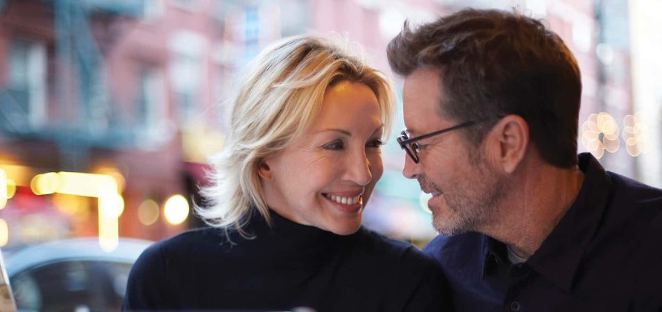 A man wearing glasses and woman in a black turtleneck shirt smile lovingly at each other.