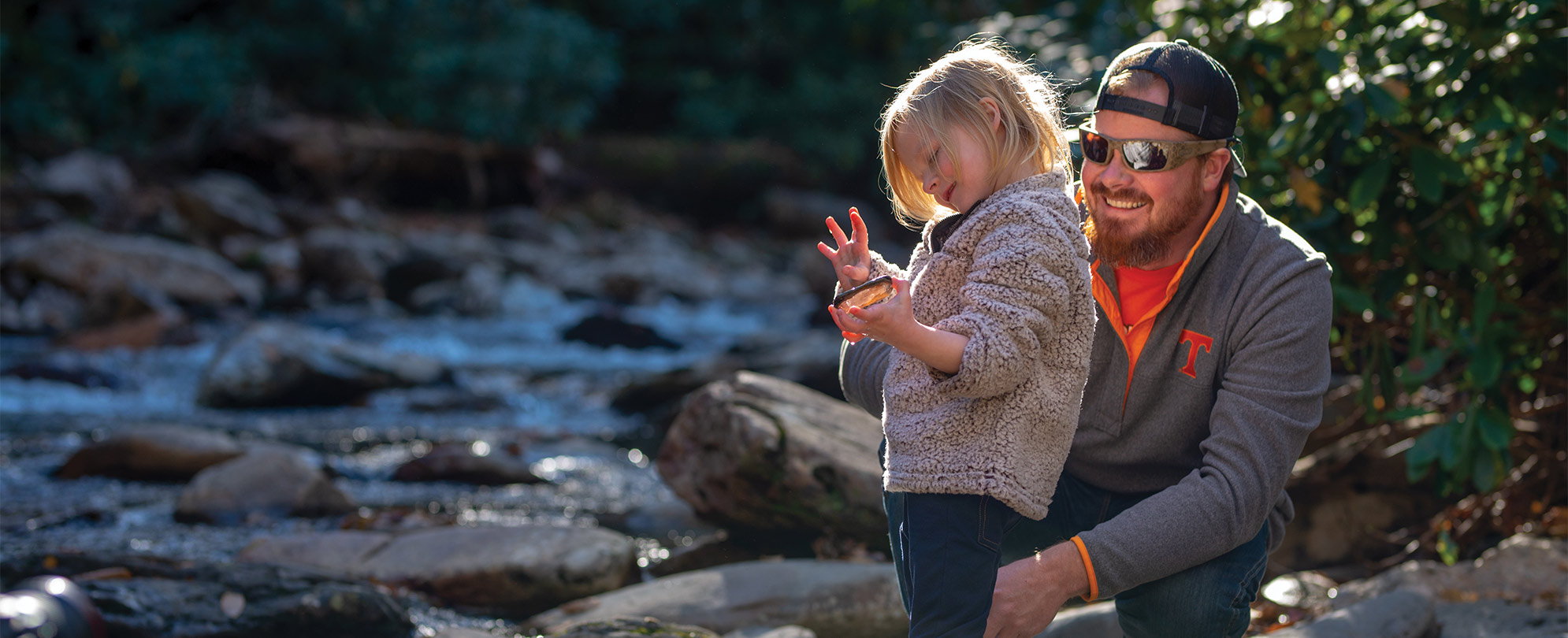 A smiling father bent down to his daughter's level as she plays with a rock down by the river